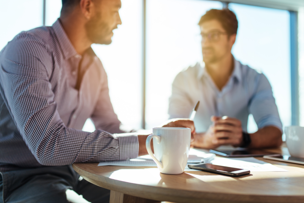 Atmospheric off-focus photo of two professional men talking in a semi-professional setting, sitting at table surrounded by documents, coffee cups, and a mobile phone, discussing cash flow lending