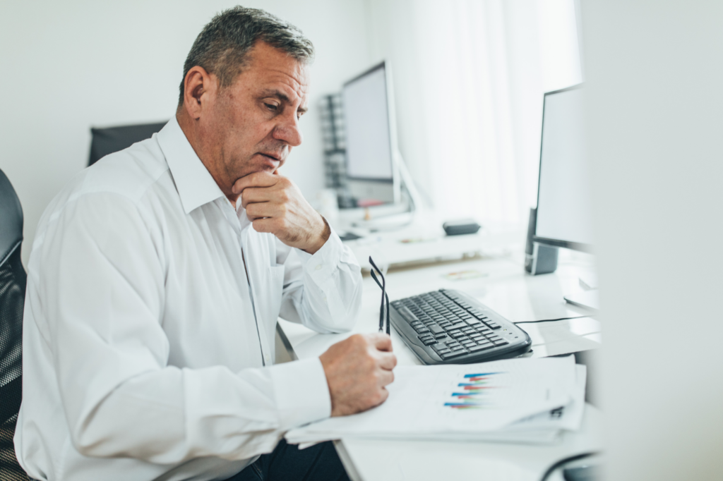 An older professional man holds his glasses in one han d and his chin in another, looking intently at documents while seated in front of a desktop computer, concept photo for assessing debts for debt consolidation