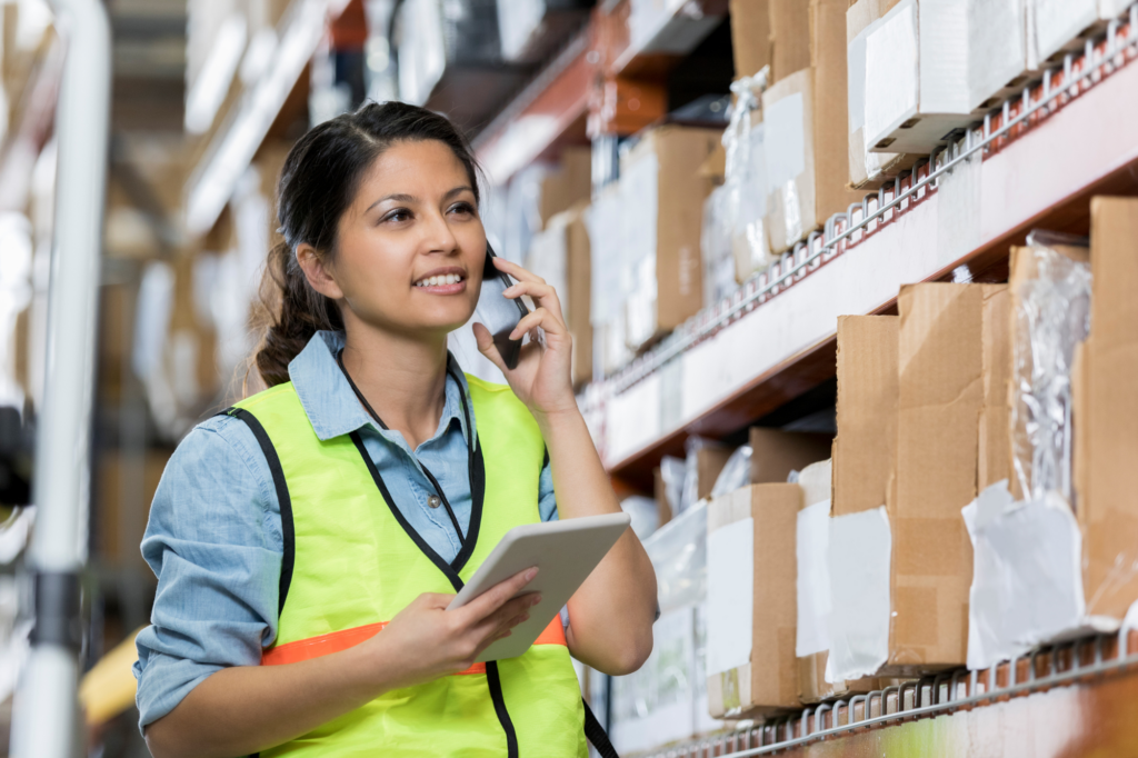 Woman in high-visibility vest smiles as she speaks on the phone and holds an electronic tablet, Talking to lenders or clients in supplying business, concept photo for getting cash flow lending approved.