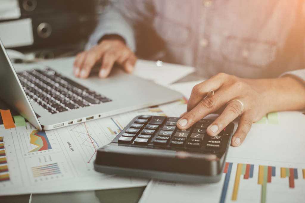 Cropped atmospheric photo of a man calculating finances, using a calculator and laptop, surrounded by documents. Concept photo for calculating projected income for cash flow lending