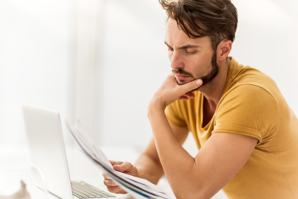 Side view of man with his hand on his chin, concentrating on reading documents while in front of laptop, concept photo for assessing debts for debt consolidation loans in Australia