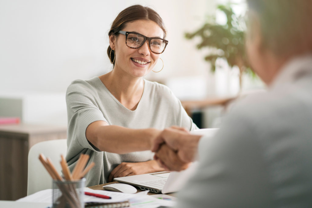 A woman in thick eyeglasses smiles as she shakes hands with a man sitting across her, concept photo for a business owner getting approved after applying for private lending in Australia