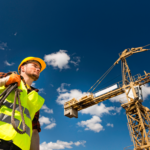 Atmospheric bottom angle photo of a construction worker walking, clear blue skies visible with construction crane in the background