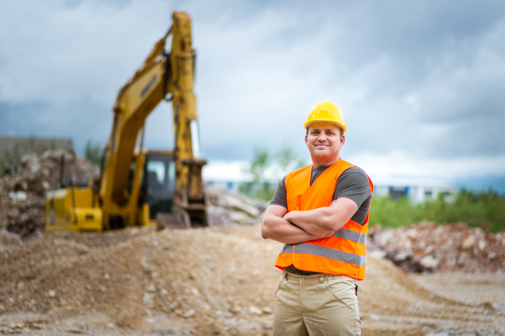 Construction worker or construction business owner smiles cheerfully with arms crossed, industrial digging equipment in the background, concept photo for getting funding for construction business