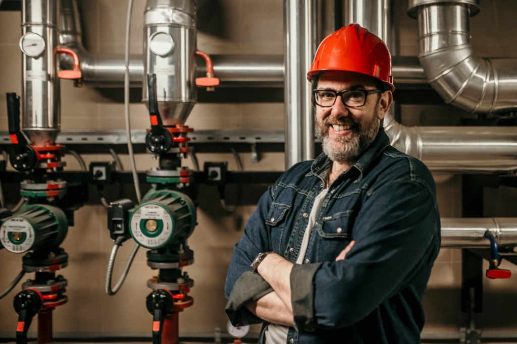 Commercial plumber with red hard hat poses in front of pipes, arms crossed, smiling at the camera