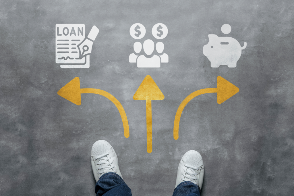 Top view of a man’s feet, white sneakers and dark jeans, visible graphic of three arrows pointing in different directions. Icons for loans, investors, and personal savings are placed on top of the arrows. Concept photo of selecting between business loans, equity financing, and bootstrapping