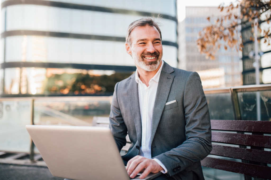 A man in a suit smiles at something off frame, sitting outdoors with a laptop, concept photo for a business owner successfully paying tax debt