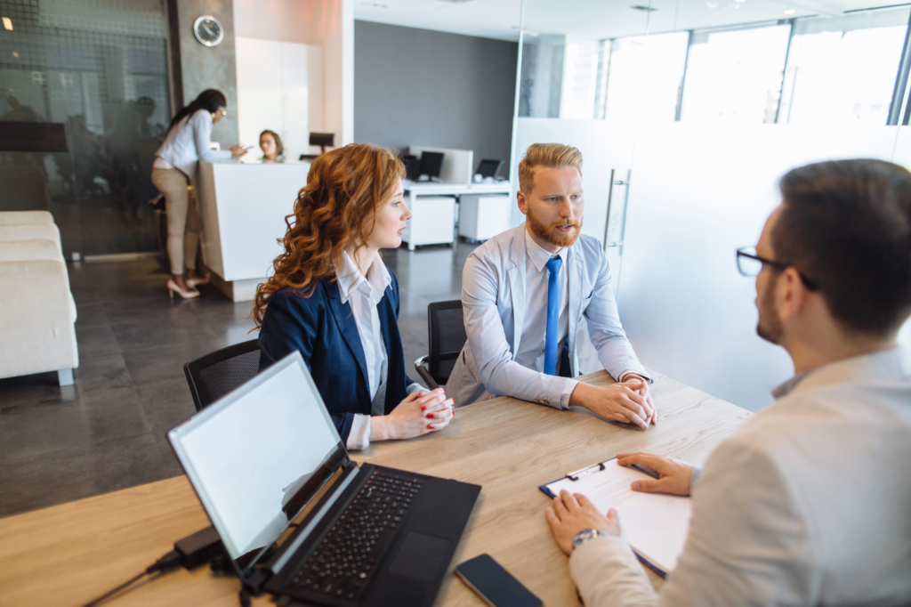 A man and woman dressed professionally sits across the table from a man holding a clipboard, concept photo for business owners discussing an unsecured business loan with a lender or loan expert