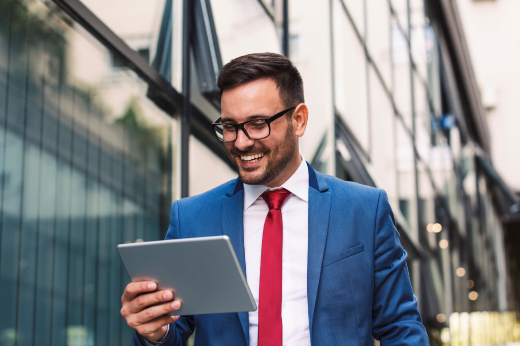 A man in a blue suit and red tie and wearing glasses cheerfully reads what’s on his electronic tablet, walking outside building with glass windows, concept photo for getting an unsecured business overdraft approved