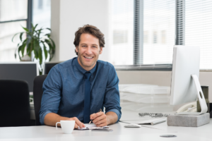 Man in a blue button-down shirt and tie sits on an office chair in front of a desk, looking directly at the camera and smiling, concept photo for business owner who got financing from a private lender