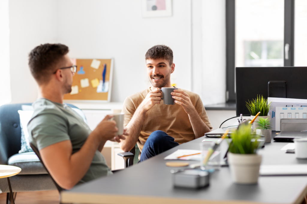 Two men drinking coffee sit side by side at a long office or conference table discussing cheerfully, concept photo of business partners discussing plans after getting funding through an overdraft facility