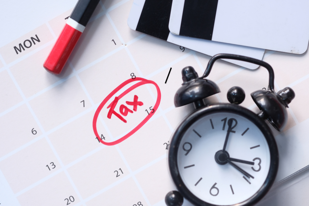 Cropped photo of a small alarm clock and calendar with a date and the word “tax” encircled in red ink. Concept photo of paying tax debt quickly before due date