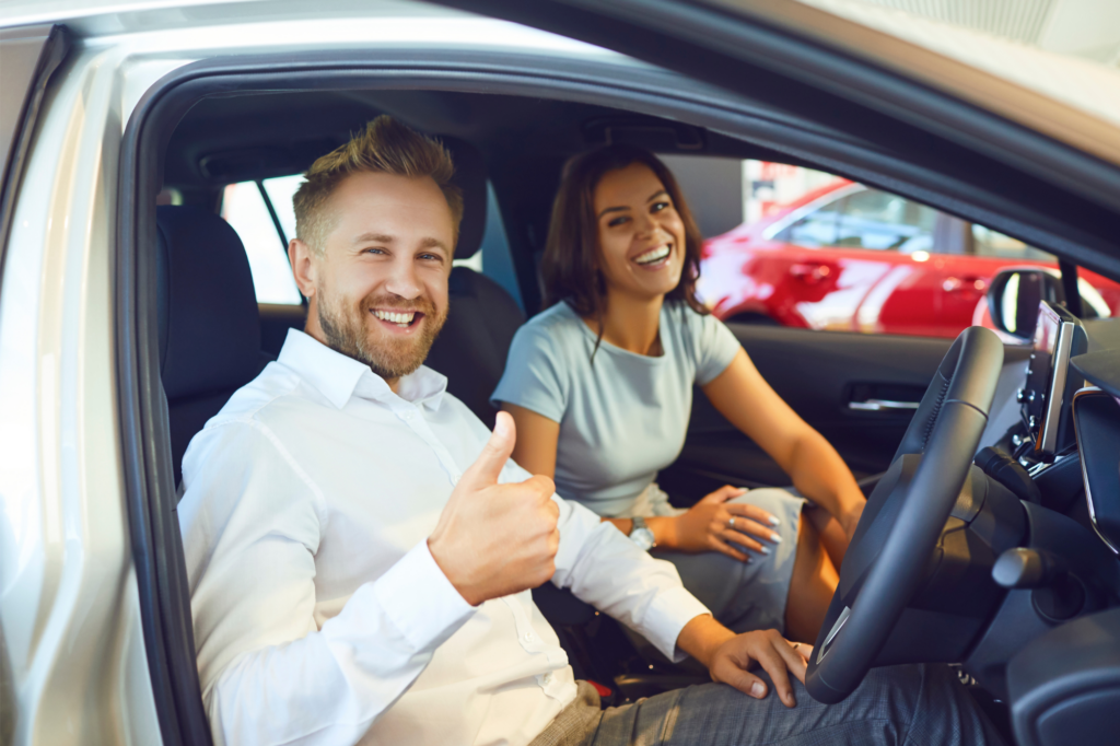 A couple sitting in a brand new car poses happily after their car loan got approved, man doing a thumbs up sign while in the driver’s seat