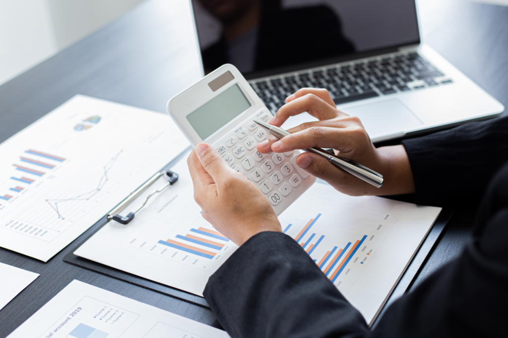 Cropped zoomed photo of a woman’s hands holding a pen and a calculator with papers and a laptop visible on the table, business owner calculating tax debt for a payment plan