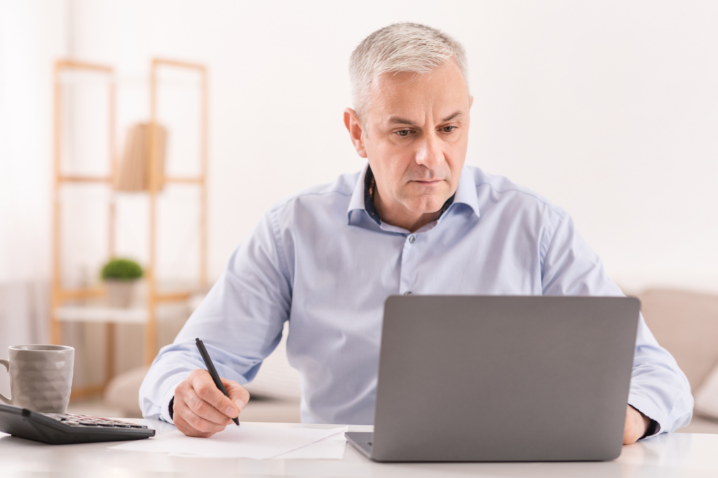 An older man concentrating, looking at his laptop while taking down notes, a business owner calculating tax and setting up a payment plan