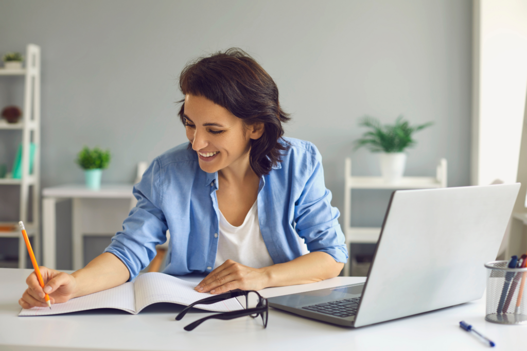 A woman cheerfully jots down something on a piece of paper, laptop visible on the table, checking requirements for SMSF loan