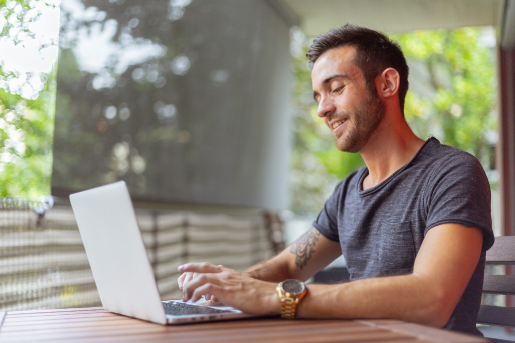 A man seated in the outdoor area of a cafe smiles as he types on his laptop, concept photo of a man successfully recycling debt