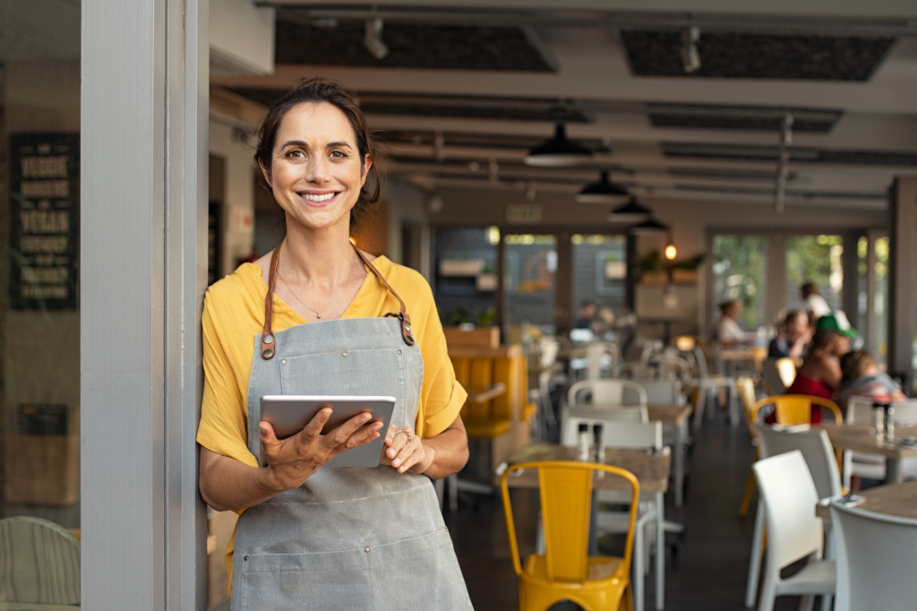 A woman in an apron holding an electronic tablet smiles while standing by the doorway of her small business cafe, concept photo for business owner choosing the right cash flow loan