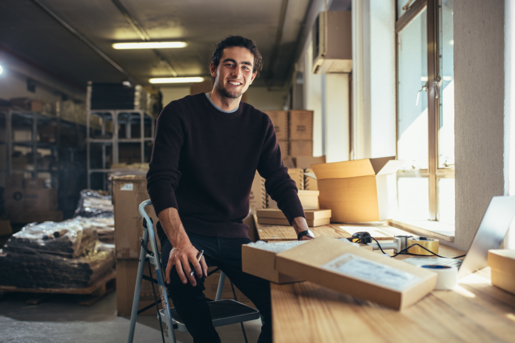 A young man sits on a stepladder inside a small business warehouse, concept photo of a young business owner securing a cash flow loan to buy inventory for his business.