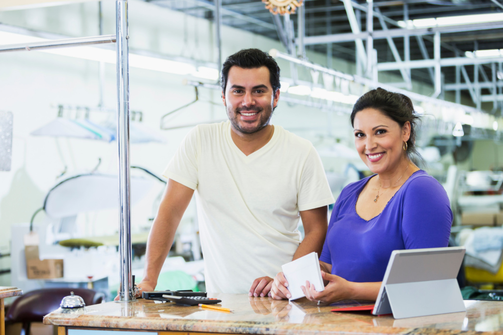 A couple smiles directly at the camera while posing at the front desk of a brightly lit business warehouse or factory, concept photo for business owners happy that they paid off daily costs with a cash flow loan