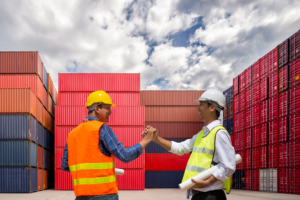 Two men in high visibility vests and hard hats shake hands in agreement, container vans in the background, concept photo for a business owner and supplier being on good terms because of import finance