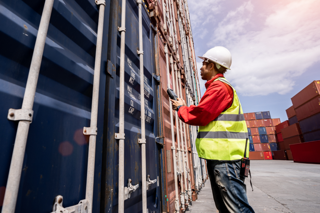 A man in a high visibility vest and a hard hat holds a device against a shipping container, a concept photo of a supplier getting materials ready for sale to an Australian business