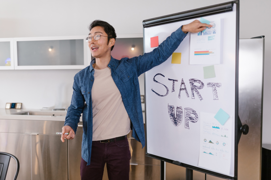 A young man in glasses points to a white board, presenting business plans, white board has the words “start up” written on it surrounded by papers, notes, and charts.