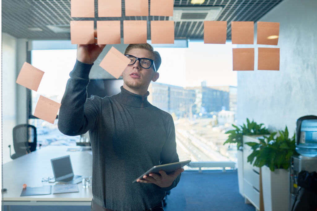 A business owner in thick black glasses holds an electronic tablet while writing on a sticky note stuck on to a clear glass panel in a conference room, business owner choosing between financing options for his startup