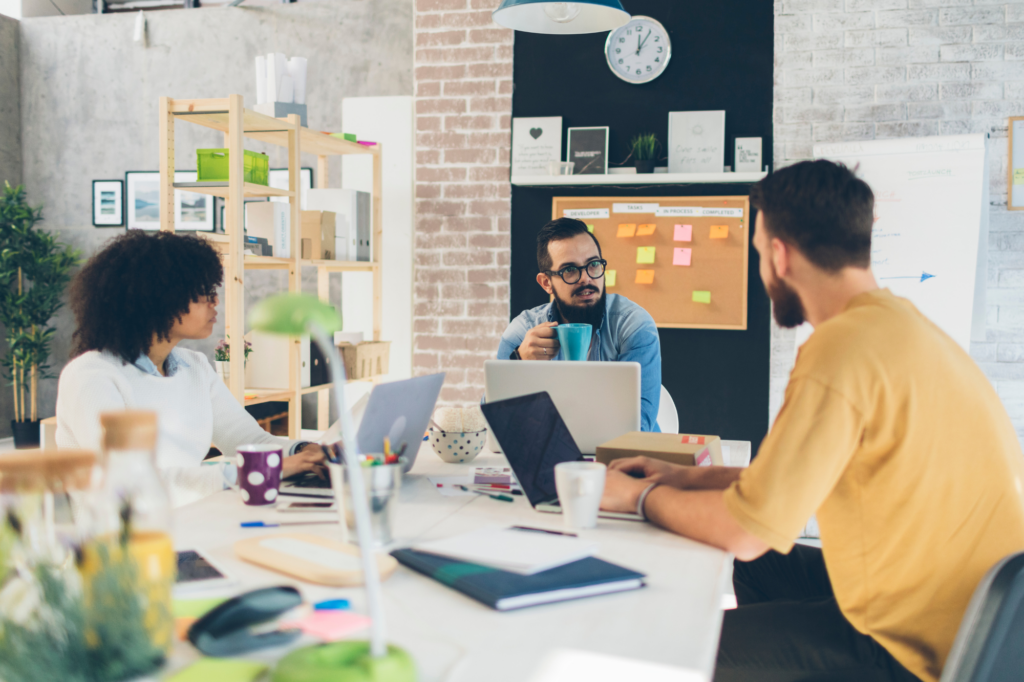 A group of young adults seated on a conference table discuss business while drinking coffee and using their laptops, business owners discussing getting a business loan for startups