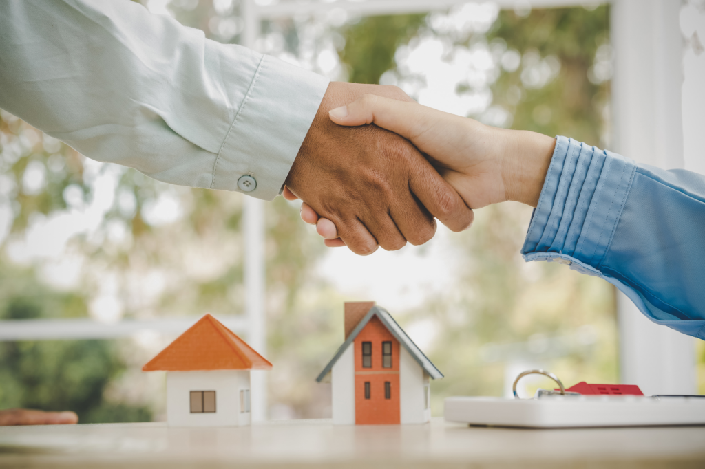 Cropped photo showing just the hands and arms of two people shaking hands, two miniature model houses, a house key, and a calculator shown in the background, concept photo for a home loan being approved