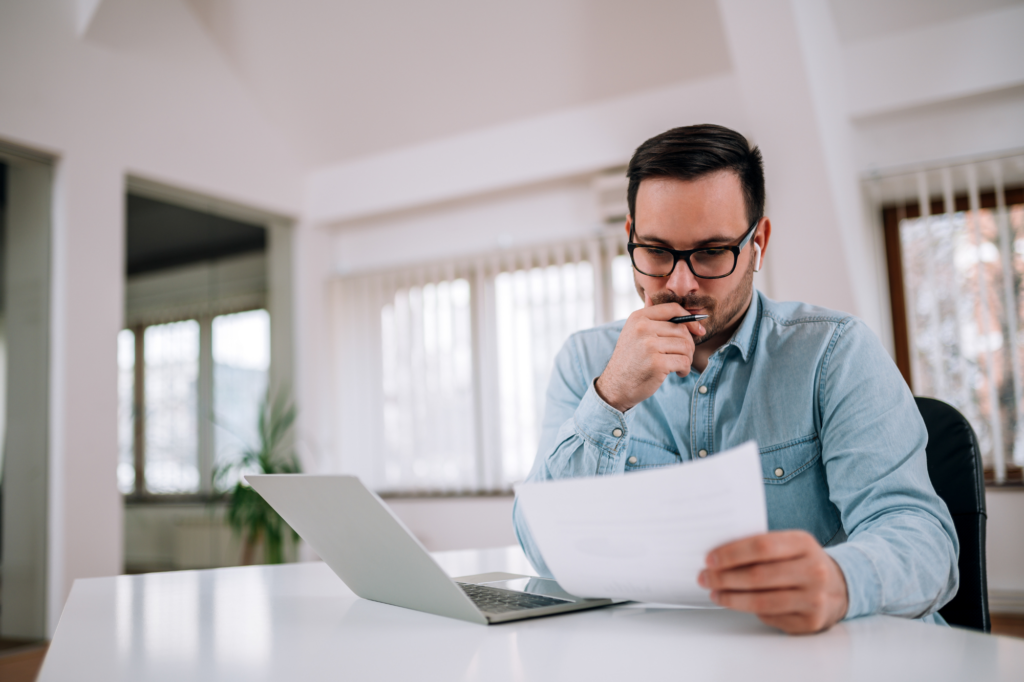 A man sits at a table with his laptop open, he is holding a document and reading it in concentration, a man considering debt recycling weighing its risks and misconceptions