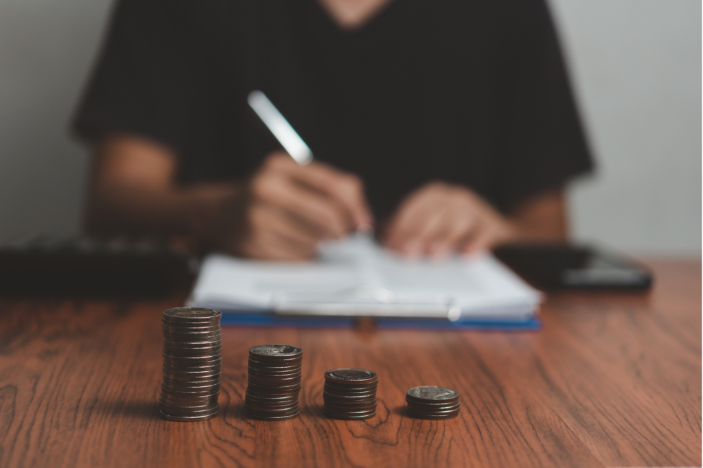 A cropped photo of a man writing on paper, with piles of coins in decreasing order in focus, concept photo for a man in debt, calculating debt