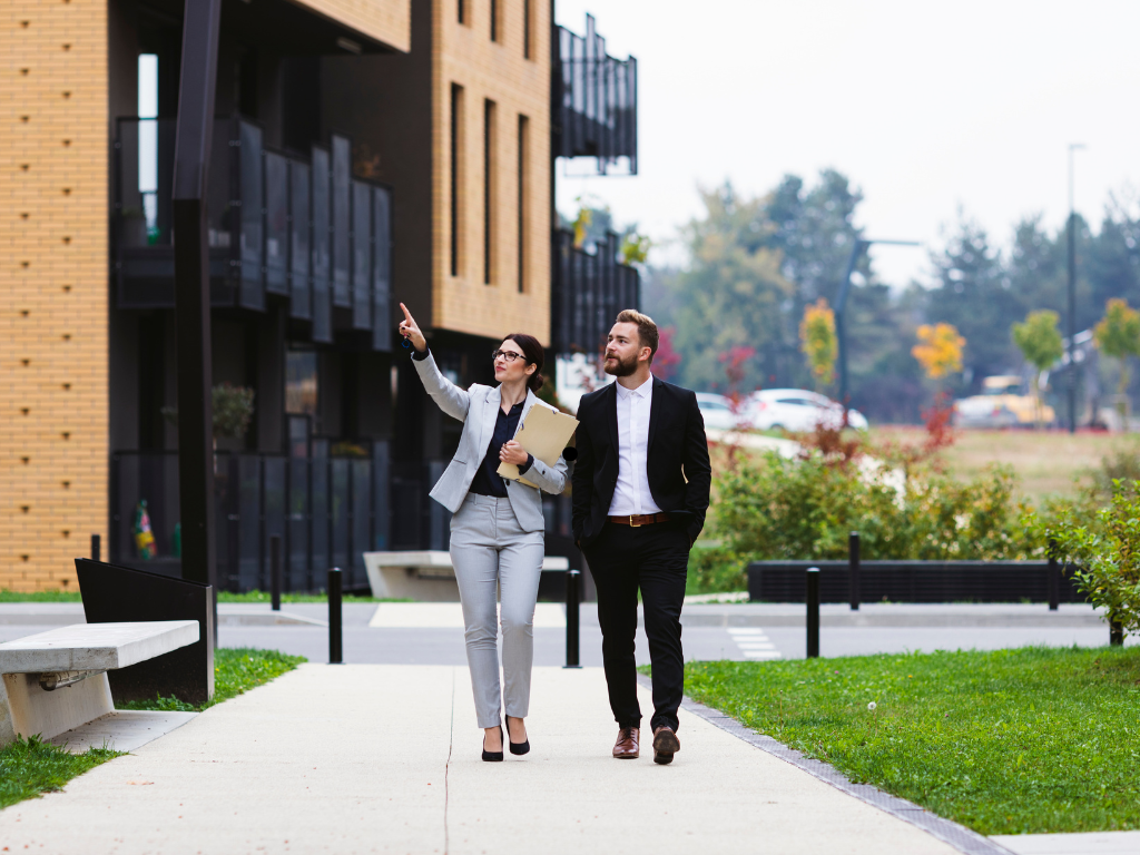 A woman points to a building while a man walking beside her looks, a realtor or seller of a commercial property presents the space to a business owner, a business owner using investment loans to purchase property