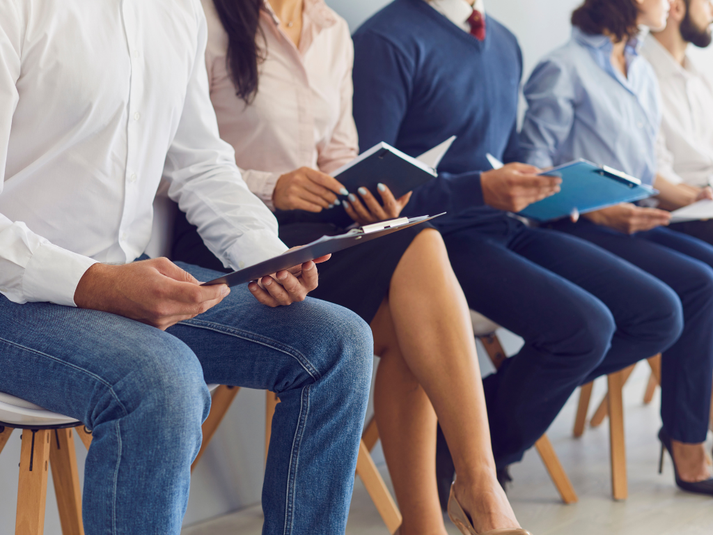 Cropped photo of several people sitting on a row of chairs against a wall, each person reading notes or files, applicants for a job opening waiting for their turn, using business investment loans to hire new personnel