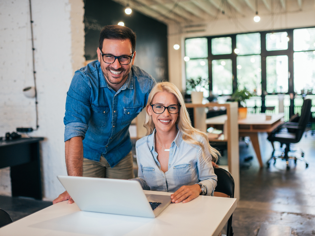 A man and a woman smile at the camera, using a laptop, business owners happily preparing for seasonal fluctuations with a business cash flow loan