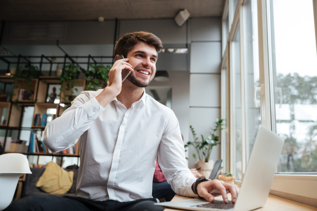 A man happily answers his cellphone as he types on his laptop with one hand, non-resident startup business director securing funding