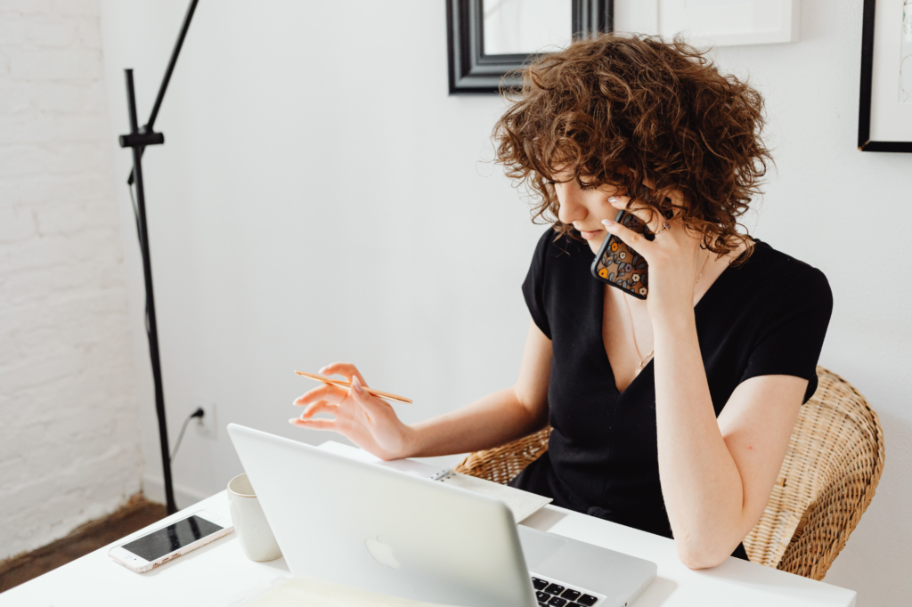 A woman speaks on the phone while looking at her laptop and taking down notes on a piece of paper, a multitasking startup business owner looking for non-resident loans