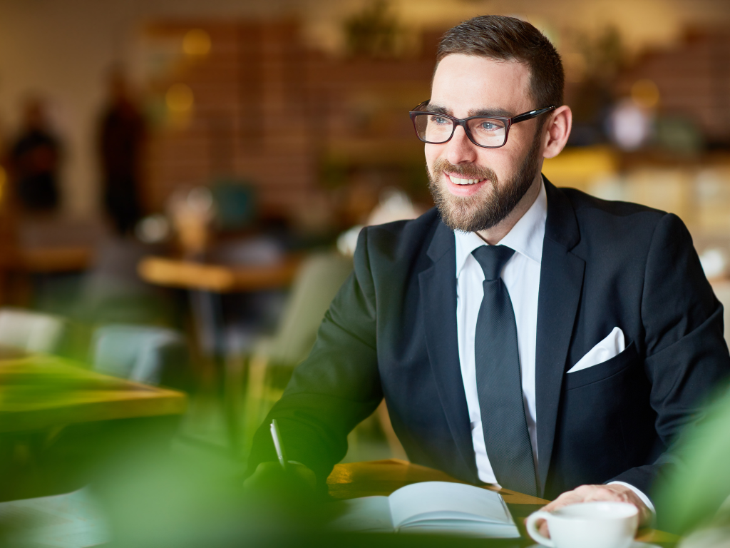 A man in a suit and glasses sits at a cafe and happily takes notes on his notebook, concept photo of a business owner considering a business line of credit for his business