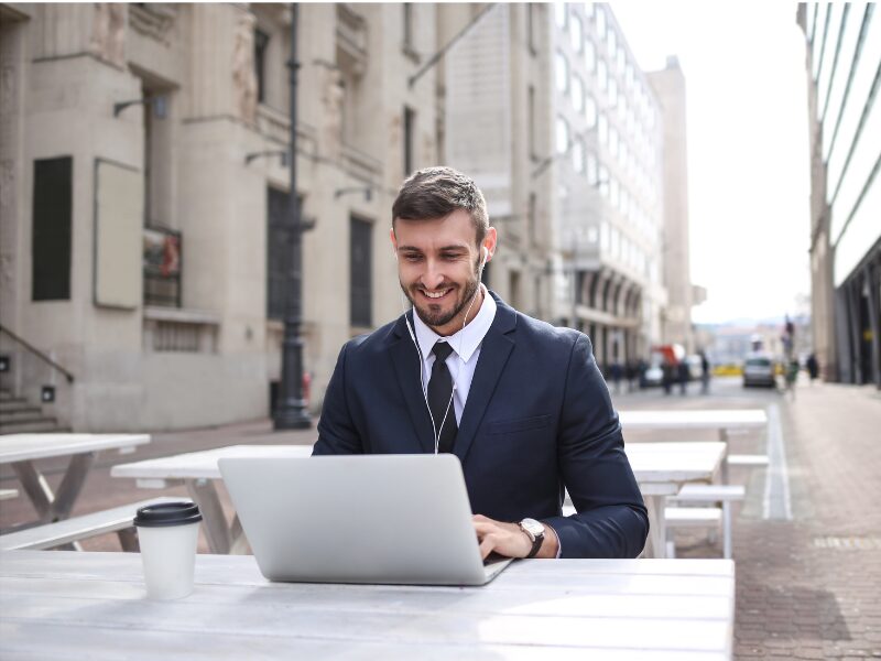 A man in a suit sits on a table on the outside of a cafe, typing on laptop, business owner enjoying the benefits of a quick business loan