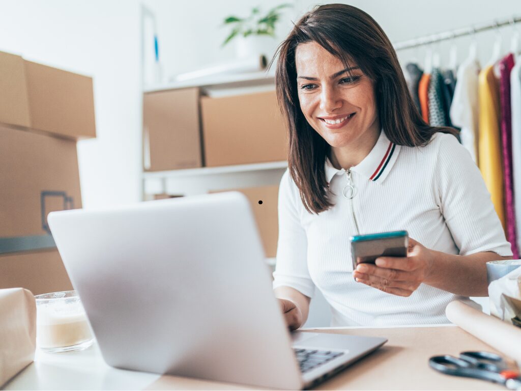 A woman smiles as she holds her cell phone while using her laptop, concept photo of a business owner securing quick business loans for her business