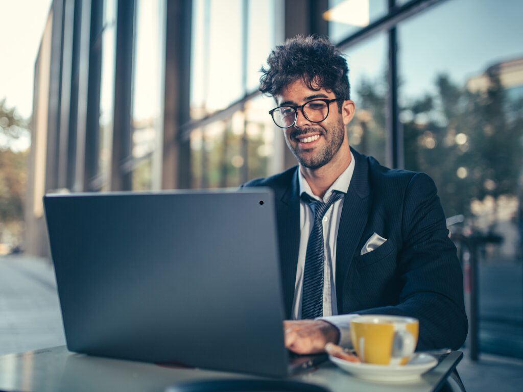 A man in a suit sits on a table on the outside of a cafe, typing on laptop, business owner enjoying the benefits of a quick business loan