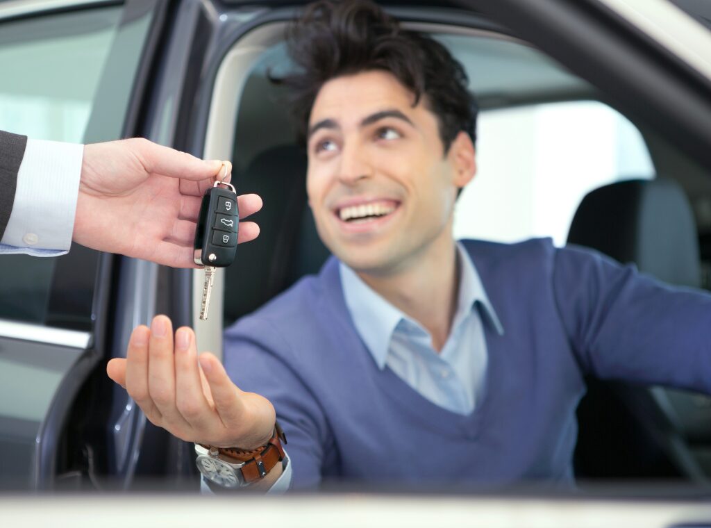 A man smiles as he sits in the driver’s seat of a new car, accepting a car key from a dealer, lifestyle creep happens when increasing discretionary spending like buying a car