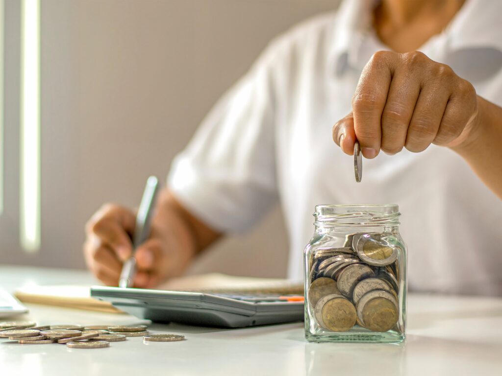 Cropped photo of a man making calculations for his savings and putting a coin inside a small coin jar, concept photo of budgeting and saving to prevent lifestyle creep