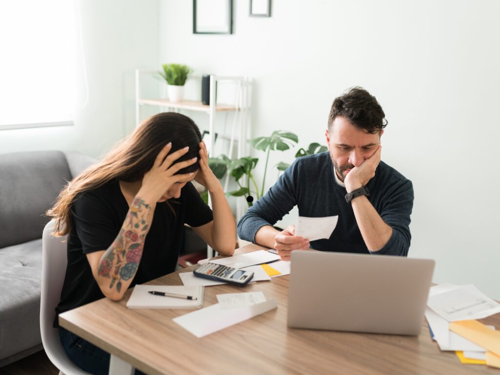 A man and a woman surrounded by scattered bills stress over finances, couple experiencing financial difficulties because of lifestyle creep