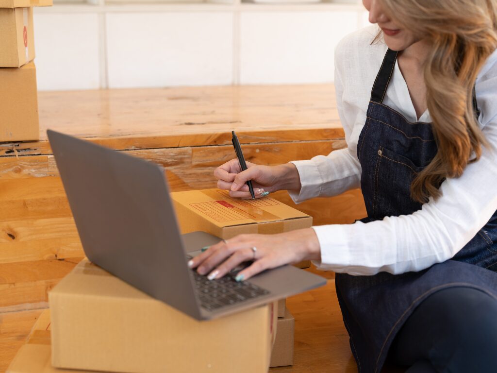 Cropped photo of a woman writing on a delivery box and using a laptop, business owner packing orders