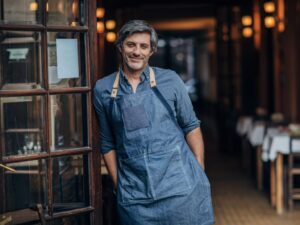 A man in a denim apron leans against the doorway of a restaurant, a happy business owner running a food establishment, using working capital for business