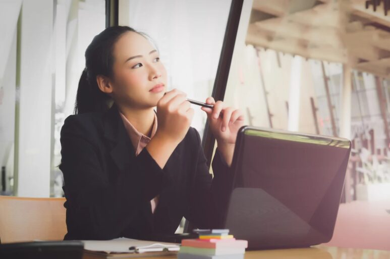 A female business owner holding a pen in front of her laptop, looking out thoughtfully while considering how to secure fitout financing in Australia for her workspace.