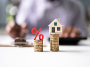 Two piles of coins, the shorter pile has a red percent sign on top while the taller pile has a miniature house on top, concept photo for an interest-only home loan