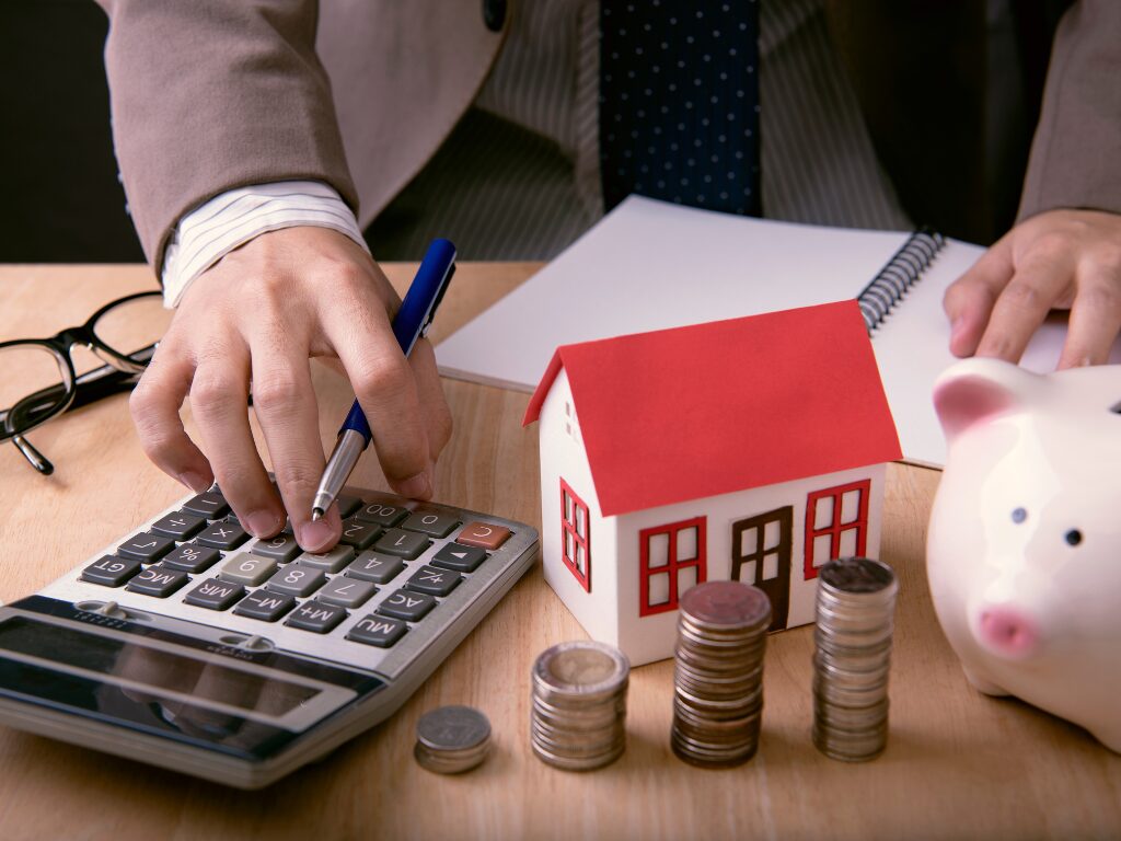 cropped photo of a man typing on a calculator while holding a pen, doing calculations, notebook, piggy bank, miniature house, and piles of coins on the table, concept photo of a man doing calculations for an interest-only loan