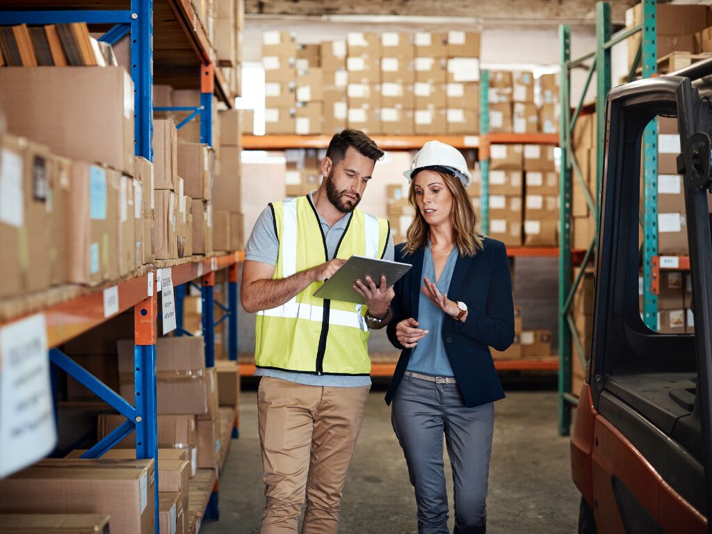 A man in a high visibility vest and a woman wearing a hard hat discuss as they walk through a warehouse, logistics company owners discussing accounts receivable financing to assist cash flow